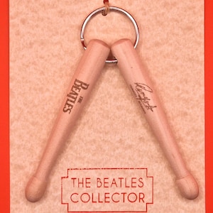 Puede incluir: Un llavero con dos baquetas de madera. Una tiene la inscripción "The Beatles", la otra "Ringo Starr". Las baquetas están unidas a un anillo plateado y se muestran en una tarjeta marrón claro con el texto "THE BEATLES COLLECTOR" en un marco rojo.