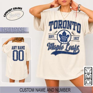 Può includere: Una t-shirt color avorio con una grafica blu effetto invecchiato del logo dei Toronto Maple Leafs, il testo TORONTO, EST. 1917 e Maple Leafs. Il retro della maglietta mostra il testo ANY NAME e il numero 00 in un carattere universitario blu.