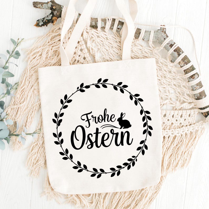 May include: A cream-colored tote bag with a black circular wreath design. The wreath surrounds the words "Frohe Ostern" and a silhouette of a rabbit. The bag has two cream-colored handles.