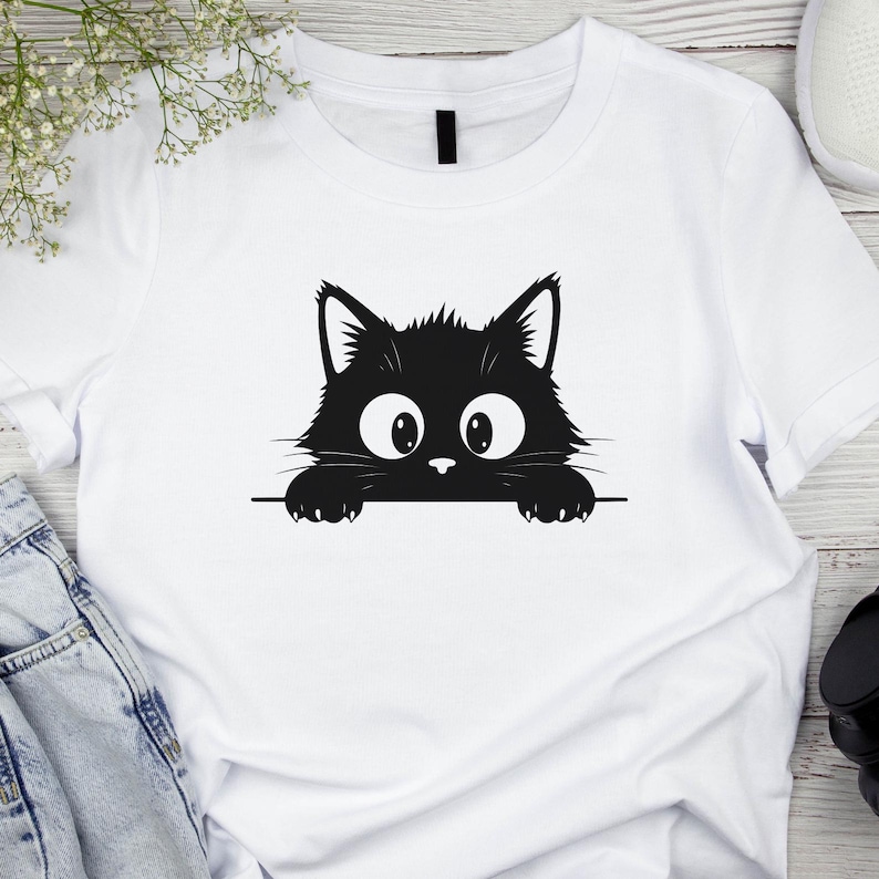 May include: White t-shirt featuring a black cat graphic. The cat has large, wide eyes and is peeking over a horizontal line. The design is simple and bold, perfect for cat lovers.