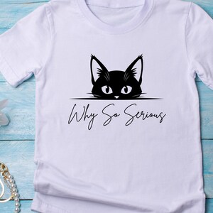 May include: White t-shirt featuring a black cat graphic with the phrase "Why So Serious" in a handwritten font. The cat's eyes and ears are visible above the text, creating a playful design.