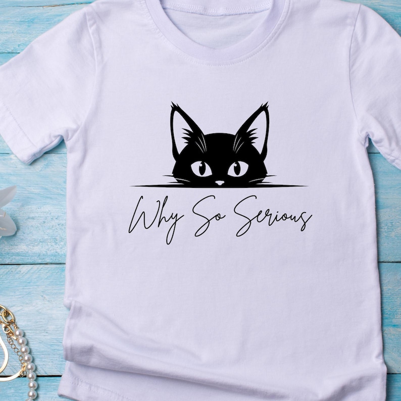 May include: White t-shirt featuring a black cat graphic with the phrase "Why So Serious" in a handwritten font. The cat's eyes and ears are visible above the text, creating a playful design.