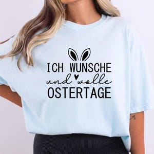 May include: A light blue t-shirt with the German text "ICH WUNSCHE und wolle OSTERTAG" in black, along with a bunny ears graphic. The shirt is a casual style.