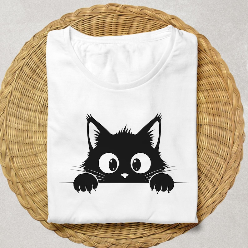 May include: White t-shirt featuring a black cat graphic. The cat's head and paws peek over a horizontal line, with large eyes and a curious expression. The shirt is folded neatly on a woven, light brown surface.
