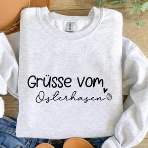 May include: A light gray sweatshirt with the text "Gr&uuml;sse vom Osterhasen" in black script. A small heart and an Easter egg graphic are next to the text. The sweatshirt is folded on a surface.