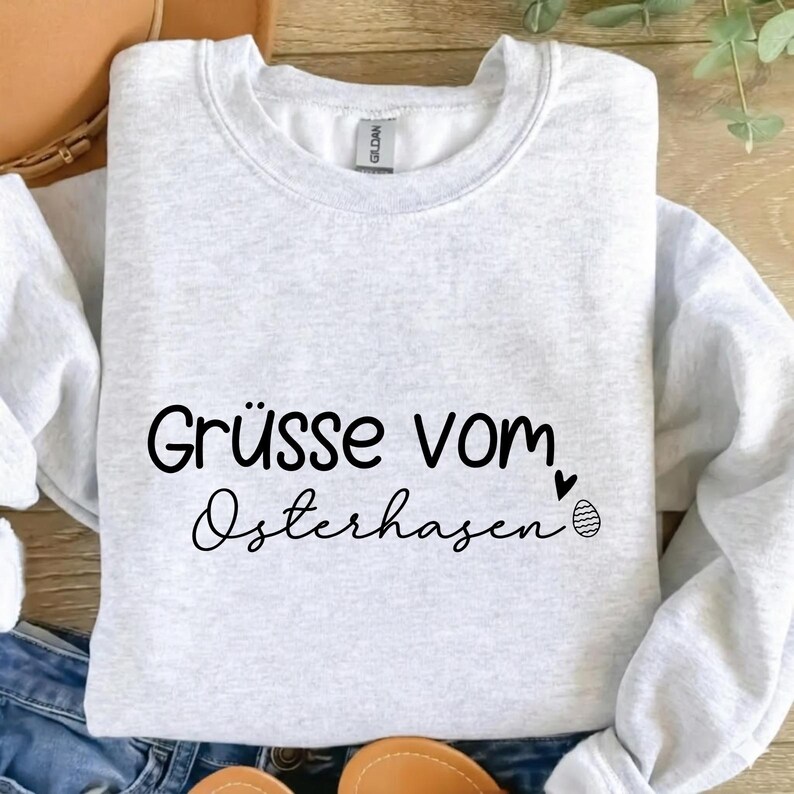 May include: A light gray sweatshirt with the text "Gr&uuml;sse vom Osterhasen" in black script. A small heart and an Easter egg graphic are next to the text. The sweatshirt is folded on a surface.