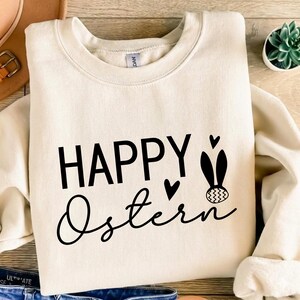 May include: Cream-colored sweatshirt with the words "HAPPY Ostern" in black script and block letters. The design includes heart shapes and a bunny with an Easter egg. The sweatshirt is folded.
