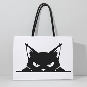 May include: White tote bag featuring a black graphic of a cat's face with pointed ears and an intense gaze. The bag has black rope handles, offering a stylish and practical accessory for carrying items.