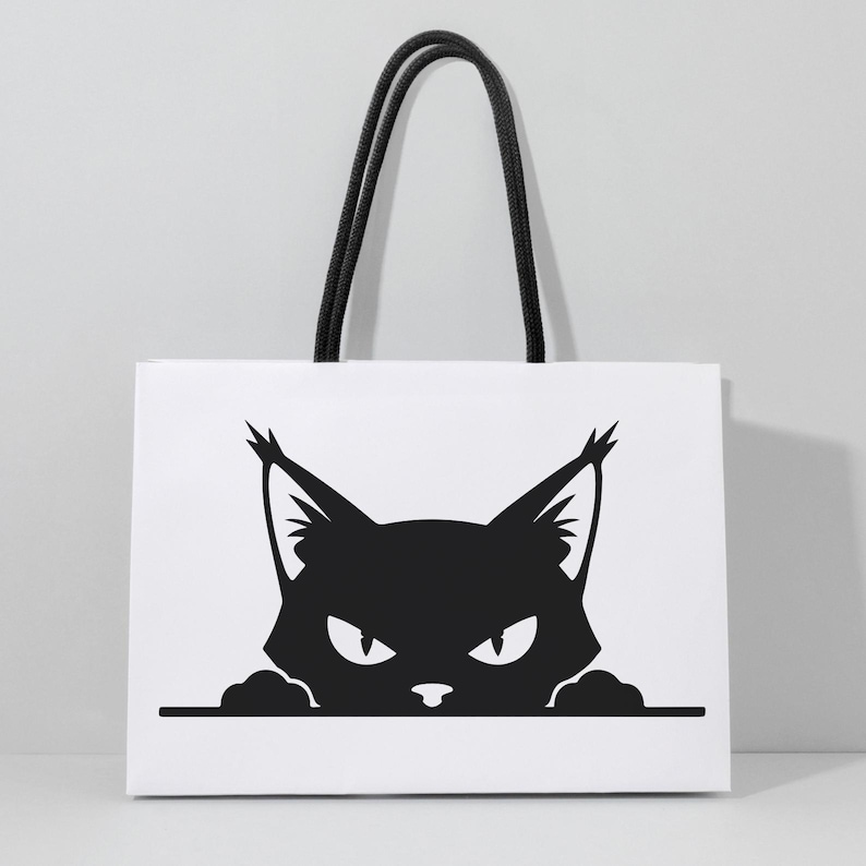 May include: White tote bag featuring a black graphic of a cat's face with pointed ears and an intense gaze. The bag has black rope handles, offering a stylish and practical accessory for carrying items.