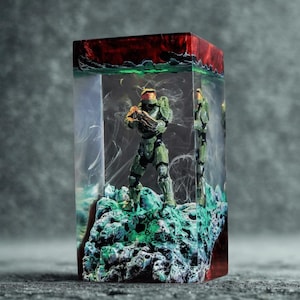 Peut inclure: Une sculpture en résine rectangulaire représentant une figurine verte et grise sur une base rocheuse. La figurine est enfermée dans de la résine transparente avec un dessus rouge et vert. La sculpture a un aspect artistique détaillé, avec un effet vaporeux et enfumé dans la résine.