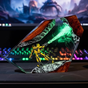 Puede incluir: Una figura de acción amarilla y negra dentro de una escultura de resina transparente. La escultura tiene una base marrón y blanca con un efecto de luz verde que emana del arma de la figura. El fondo está borroso, mostrando un monitor de computadora.