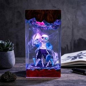 Könnte beinhalten: Harzskulptur mit einer Figur aus dem Videospiel Undertale, eingebettet in einen transparenten Block. Die Figur trägt eine blaue Jacke und dunkle Hosen, mit einem schädelartigen Kopf und einem leuchtenden blauen Auge. Der Sockel ist rot, mit blauen und violetten Akzenten.