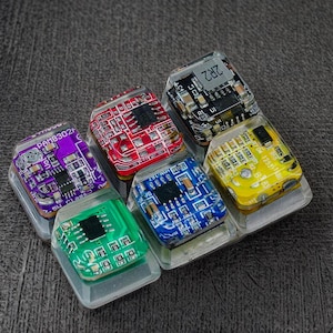 May include: A set of six colorful, custom keyboard keycaps. Each keycap features a unique circuit board design in various colors, including purple, red, green, blue, and yellow. The keycaps are encased in clear resin.