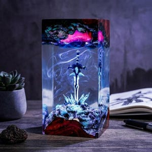 May include: A rectangular resin art piece featuring a miniature sword. The clear resin encases a detailed sword atop a colorful base of red, blue, and purple hues. The top of the resin block has a mix of pink, blue, and green colors.