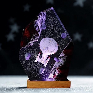 May include: A resin sculpture with a spaceship design, set on a wooden base. The spaceship is white with red details, within a dark, space-themed setting with purple and red hues. The sculpture is lit with a purple light.
