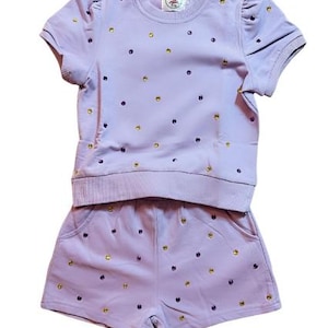 May include: A lilac-colored two-piece outfit for children, featuring a short-sleeved top and matching shorts. The set is adorned with scattered gold and purple embellishments. The top has a ribbed hem and the shorts have pockets.