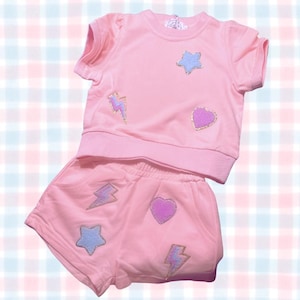 Peut inclure: Ensemble deux pièces rose clair pour enfant, composé d'un haut à manches courtes et d'un short assorti. Les deux pièces sont ornées d'écussons brodés en forme d'étoile, de cœur et d'éclair. Fond à carreaux pastel.
