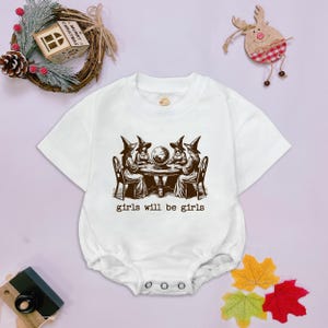Puede incluir: Un body blanco de manga corta para beb&eacute; con un estampado gr&aacute;fico marr&oacute;n de cuatro brujas sentadas alrededor de una mesa con una bola de cristal. El texto "girls will be girls" est&aacute; impreso en color marr&oacute;n debajo de la ilustraci&oacute;n.
