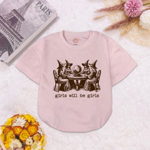 Puede incluir: Una camiseta de manga corta de color rosa claro con un estampado gr&aacute;fico marr&oacute;n oscuro de cuatro figuras con sombreros puntiagudos sentadas alrededor de una mesa con una bola de cristal. Debajo de la ilustraci&oacute;n, el texto dice "girls will be girls".