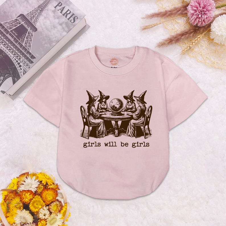 Puede incluir: Una camiseta de manga corta de color rosa claro con un estampado gr&aacute;fico marr&oacute;n oscuro de cuatro figuras con sombreros puntiagudos sentadas alrededor de una mesa con una bola de cristal. Debajo de la ilustraci&oacute;n, el texto dice "girls will be girls".