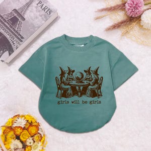 Puede incluir: Una camiseta de manga corta color verde azulado con un gr&aacute;fico marr&oacute;n oscuro de cuatro figuras con sombreros puntiagudos sentadas alrededor de una mesa con una bola de cristal. El texto "girls will be girls" aparece impreso debajo.