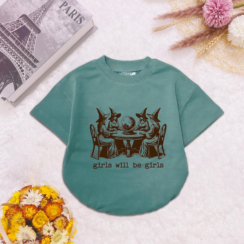 Puede incluir: Una camiseta de manga corta color verde azulado con un gr&aacute;fico marr&oacute;n oscuro de cuatro figuras con sombreros puntiagudos sentadas alrededor de una mesa con una bola de cristal. El texto "girls will be girls" aparece impreso debajo.