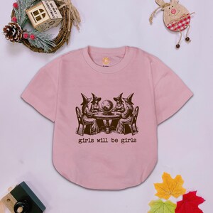 Puede incluir: Una camiseta de manga corta de color rosa claro con un estampado gr&aacute;fico marr&oacute;n oscuro de cuatro figuras con sombreros de bruja sentadas alrededor de una mesa con una bola de cristal. Debajo, el texto dice girls will be girls en letra de m&aacute;quina.