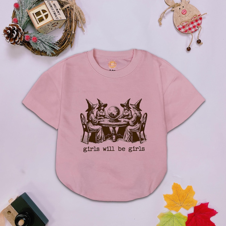 Puede incluir: Una camiseta de manga corta de color rosa claro con un estampado gr&aacute;fico marr&oacute;n oscuro de cuatro figuras con sombreros de bruja sentadas alrededor de una mesa con una bola de cristal. Debajo, el texto dice girls will be girls en letra de m&aacute;quina.
