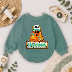 May include: A sage green long-sleeve sweatshirt featuring a graphic of an orange and yellow checkered ghost wearing a teal and white baseball cap with a bat icon. Below the ghost is a rectangular orange frame containing the name Hudson in teal.