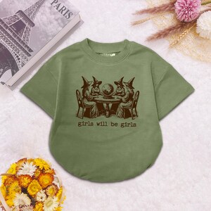Puede incluir: Una camiseta de manga corta color verde oliva con un gr&aacute;fico marr&oacute;n oscuro de cuatro brujas sentadas alrededor de una mesa con una bola de cristal. El texto "girls will be girls" est&aacute; impreso en marr&oacute;n oscuro bajo la ilustraci&oacute;n.