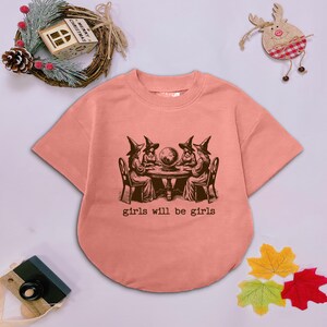 Puede incluir: Una camiseta de manga corta color melocot&oacute;n con un gr&aacute;fico marr&oacute;n oscuro de cuatro figuras con sombreros puntiagudos sentadas alrededor de una mesa con una bola de cristal. Debajo del gr&aacute;fico, el texto dice girls will be girls.