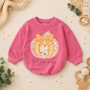 May include: A long-sleeve pink sweatshirt featuring a central graphic of a pumpkin with a large orange polka-dot bow. The pumpkin has a floral and gingham pattern with a white lace border. The name Olivia is printed in black script below the pumpkin.