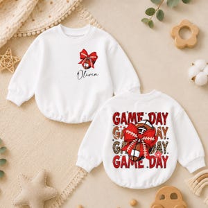 May include: A white long-sleeve sweatshirt featuring a small red bow and football graphic with the name Olivia on the front, and a large graphic on the back with the text Game Day, a football, a red bow, and leopard print accents.