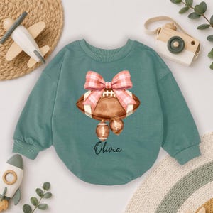 Op de afbeelding: Een saliegroen sweatshirt met lange mouwen en een centrale afbeelding van een bruine football versierd met een grote roze en wit geruite strik. Twee kleinere footballs hangen onder het hoofdontwerp en de naam Olivia staat in zwarte letters.