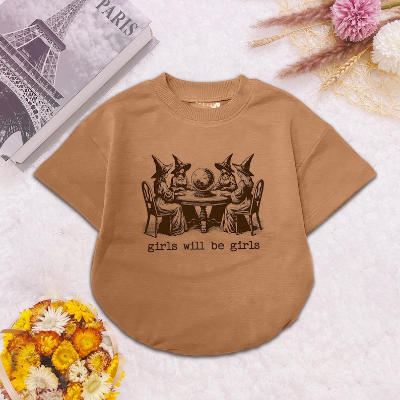 Puede incluir: Una camiseta de manga corta color canela con un gr&aacute;fico marr&oacute;n oscuro de cuatro figuras con sombreros puntiagudos sentadas alrededor de una mesa con una bola de cristal. El texto "girls will be girls" aparece impreso debajo del dibujo.