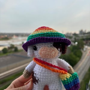Può includere: Una statuetta di mucca all'uncinetto fatta a mano, con un cappello arcobaleno e una borsa a tracolla abbinata. La mucca è bianca con macchie marroni e un naso color pesca. Il cappello e la borsa presentano vivaci strisce arcobaleno.