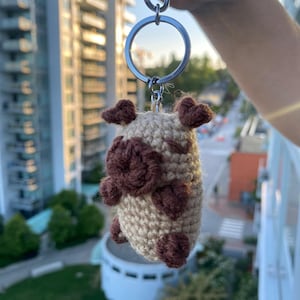 May include: A crocheted keychain featuring a brown and tan cow design. The cow has brown spots and a silver keyring. The keychain is held up against a background of buildings and trees.