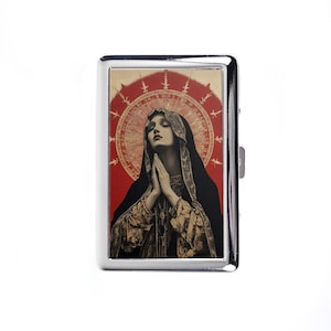 May include: A silver cigarette case with a religious image. The image shows a woman in a black veil with hands clasped in prayer, set against a red background with a golden halo.