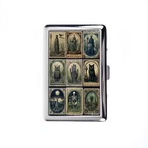 Op de afbeelding: Een zilveren metalen sigarettenetui met een verzameling vintage gotische illustraties. De afbeeldingen omvatten een uil, een zwarte kat, een hand en andere mystieke figuren. De etui heeft een rechthoekige vorm met een sluiting.