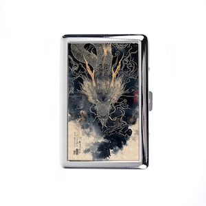 May include: A silver cigarette case with a detailed illustration of a golden dragon against a dark, cloudy background. The dragon's face is centered, with intricate details. The case has a rectangular shape and a secure clasp.