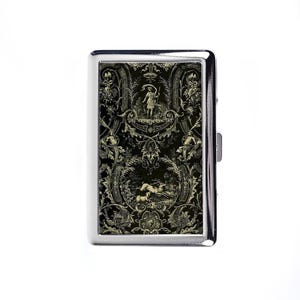 May include: A silver cigarette case with a black and cream decorative design. The design features a pastoral scene with figures, deer, and ornate floral and scrollwork elements. The case has a rectangular shape and a hinged closure.
