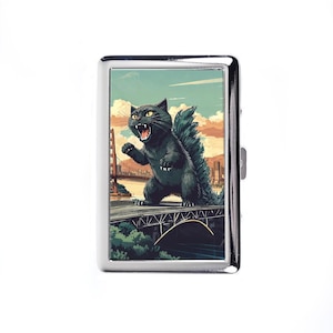 May include: A silver metal cigarette case featuring a cartoon illustration of a black cat resembling Godzilla, standing on a bridge with the Golden Gate Bridge in the background. The cat is roaring with its mouth open.