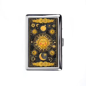 Pode incluir: Um porta-cigarros de metal prateado com um design celestial. A caixa apresenta um fundo preto com ilustrações douradas do sol, da lua, das estrelas e outros corpos celestes. O sol tem um rosto com bochechas rosadas.