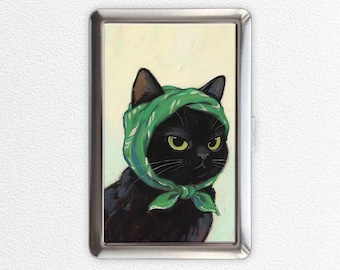 Pode incluir: Um porta-cigarros de metal prateado com uma pintura de um gato preto usando um lenço verde. O gato tem olhos amarelos e uma expressão ligeiramente carrancuda. O fundo é amarelo pálido.
