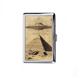 May include: A silver cigarette case featuring a sepia-toned illustration. The design depicts a pyramid below a schematic of a flying saucer. The case has a rectangular shape with a shiny, metallic finish and a side clasp.