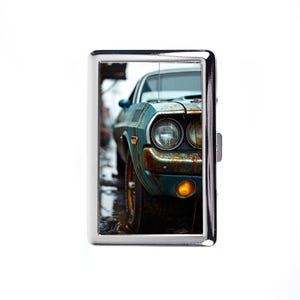 May include: A silver metal cigarette case featuring a teal vintage car with rust details. The car's headlights and front are visible, set against a blurred background. The case has a rectangular shape with a hinged opening.