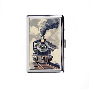 May include: A silver cigarette case featuring a vintage train illustration. The train is black with a yellow headlight and the word "LUCKY" on the front. The background shows a cloudy sky and train tracks.