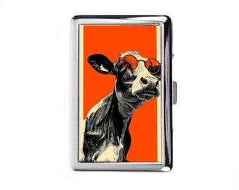 Puede incluir: Carcasa de cigarrillos de metal plateado con una ilustración en blanco y negro de una vaca con gafas de sol naranjas sobre un fondo naranja. La funda es rectangular y tiene una apertura con bisagras.