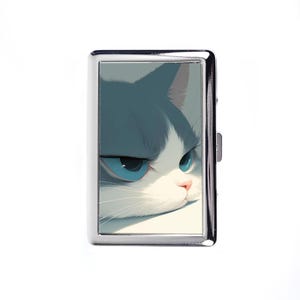 May include: A silver cigarette case featuring a close-up illustration of a cat's face. The cat has blue eyes, white fur, and gray markings. The case has a rectangular shape and a hinged lid.