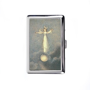 Pode incluir: Um porta-cigarros prateado com uma ilustração de fantasia. A imagem mostra uma fada com uma lua crescente e asas, em pé sobre um orbe brilhante, com estrelas e nuvens. O porta-cigarros tem um acabamento metálico brilhante.